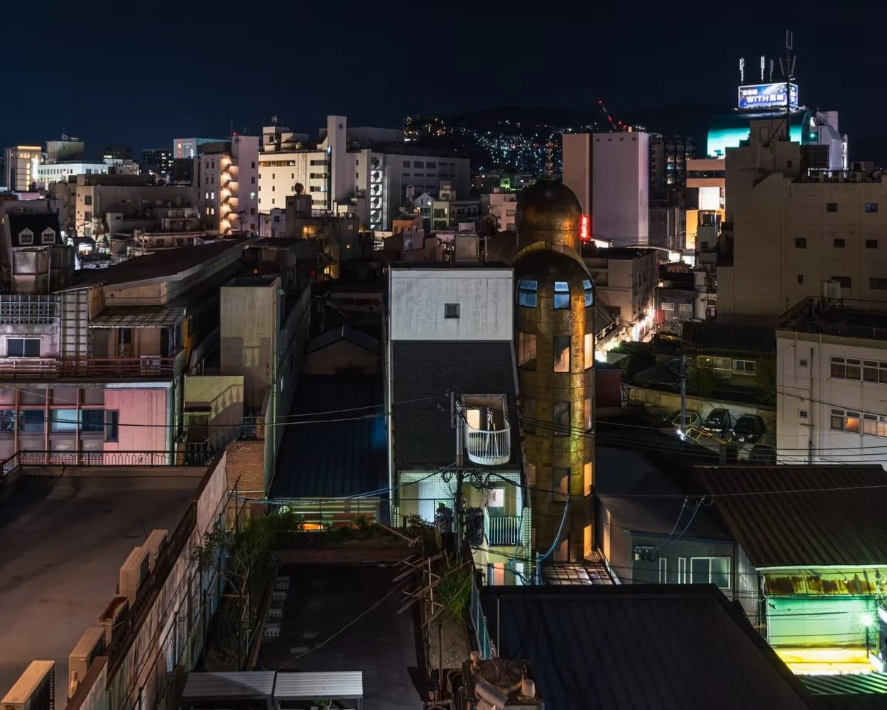 Nighttime cityscape with illuminated buildings, a golden dome-shaped tower, and a hillside in the background.