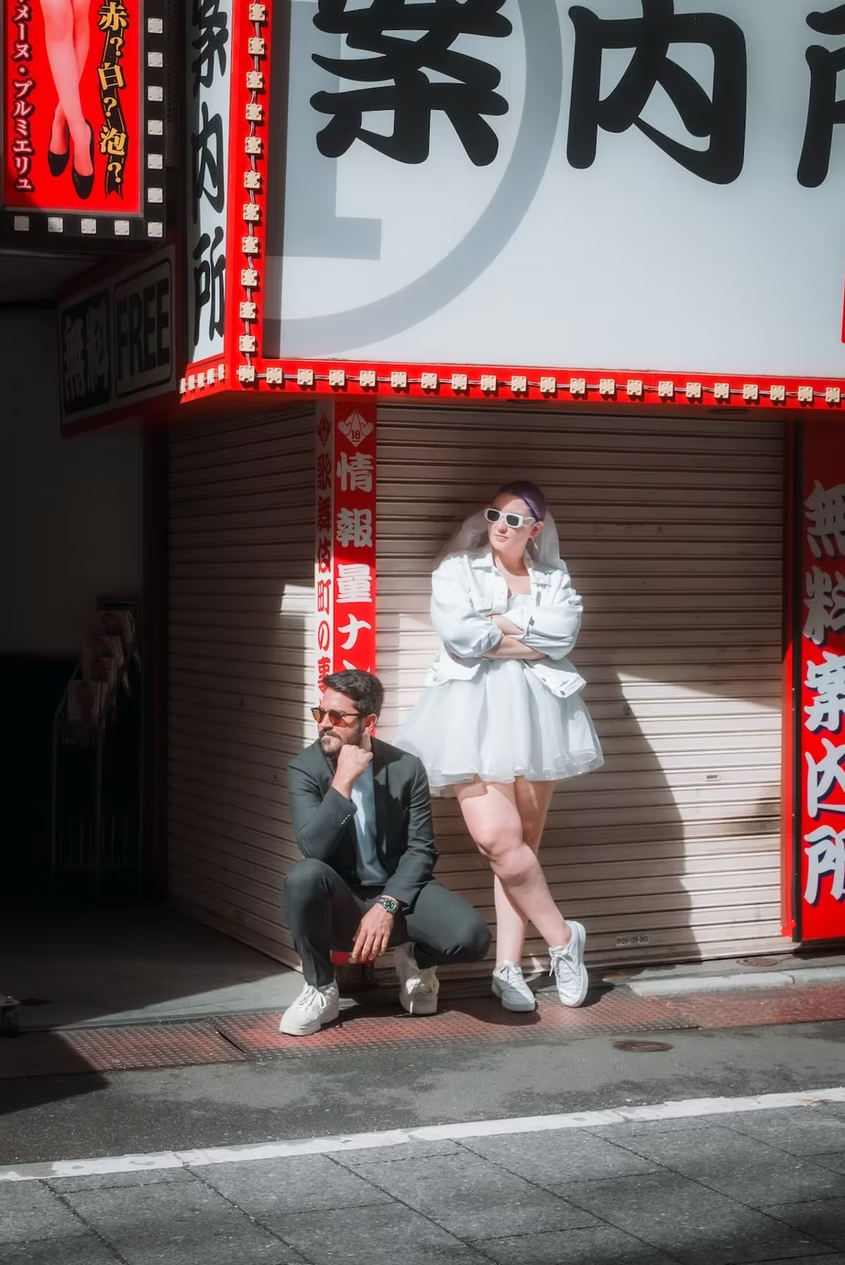 A couple sits in front of a traditional Japanese restaurant, with one person squatting and the other sitting on the ground. The background features a large neon sign with Japanese characters.