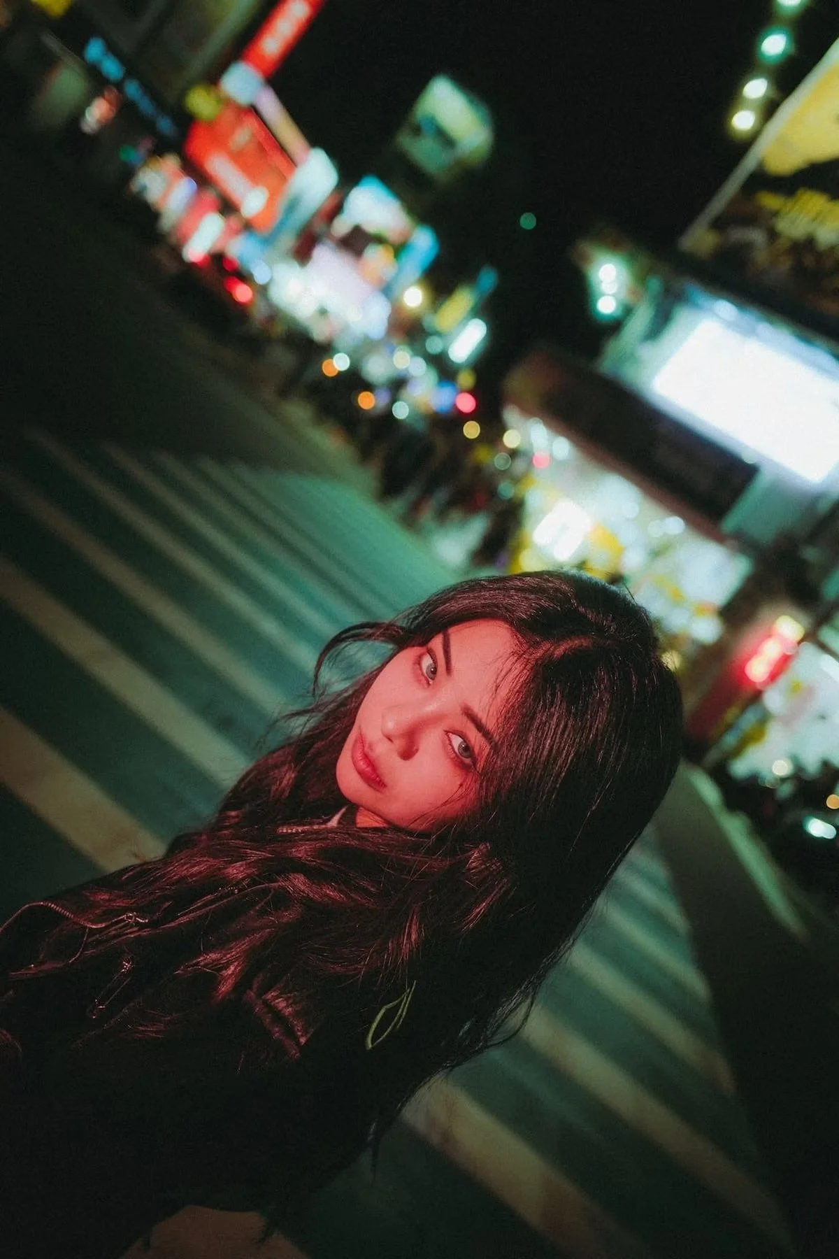 A young woman with long dark hair and a subtle smile is standing in front of a blurred city street at night, illuminated by colorful neon lights in Taipei shot by Tokyo portrait photographer Deniz Demir.