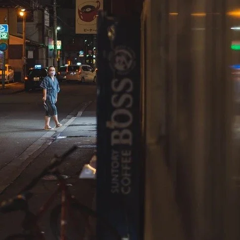 A man walks barefoot on a dimly lit Tokyo street at night, passing a blue Boss Coffee vending machine, with blurred lights of a passing bus nearby.