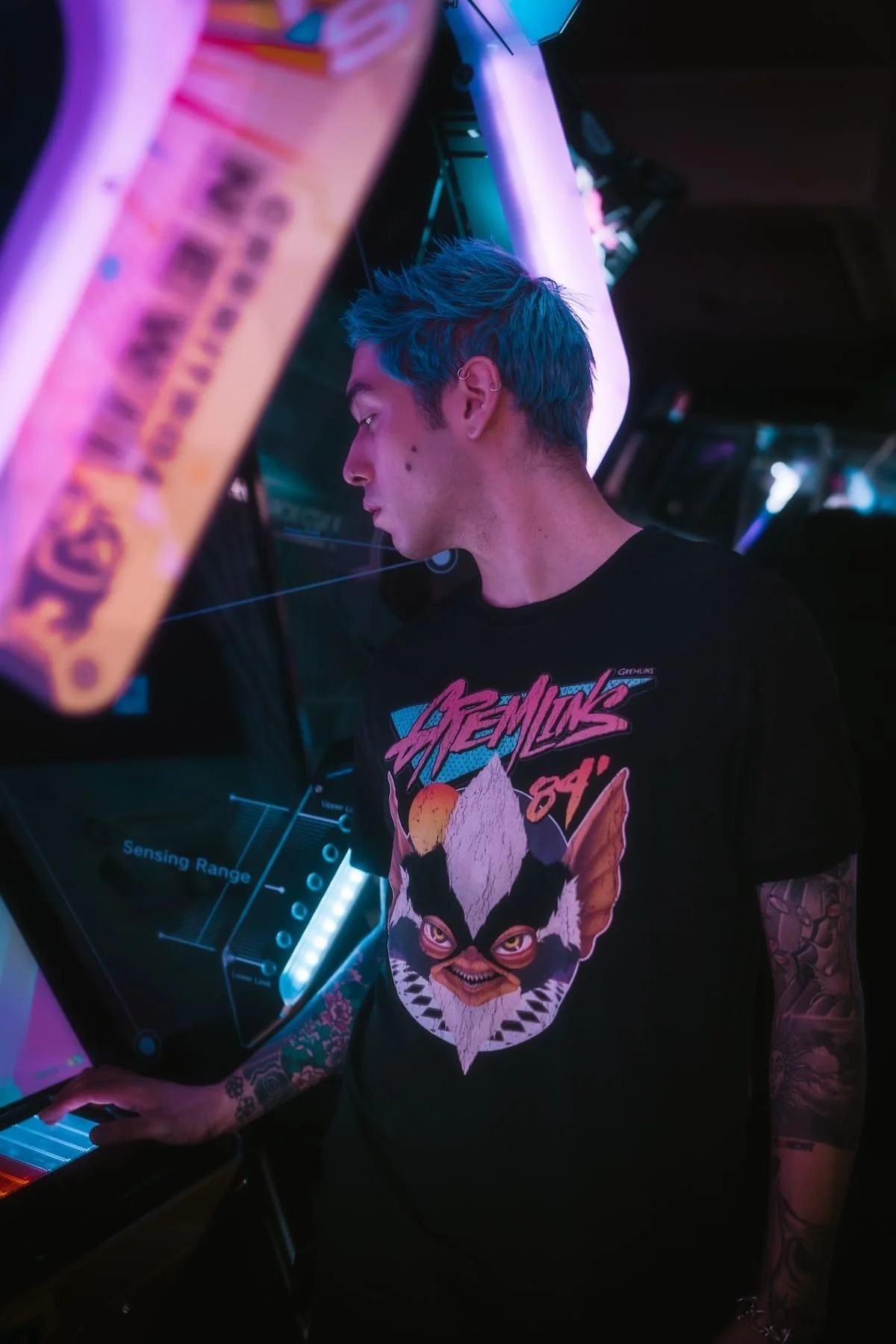 A young man with blue hair playing arcade pinball machine, wearing a black T-shirt featuring a stylized feline face and the text 'Archimic 94'.