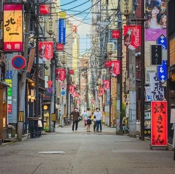 A narrow street in Tokyo lined with restaurants and bars, featuring bright red lanterns, neon signs, and overhead power lines. Pedestrians stroll ahead.