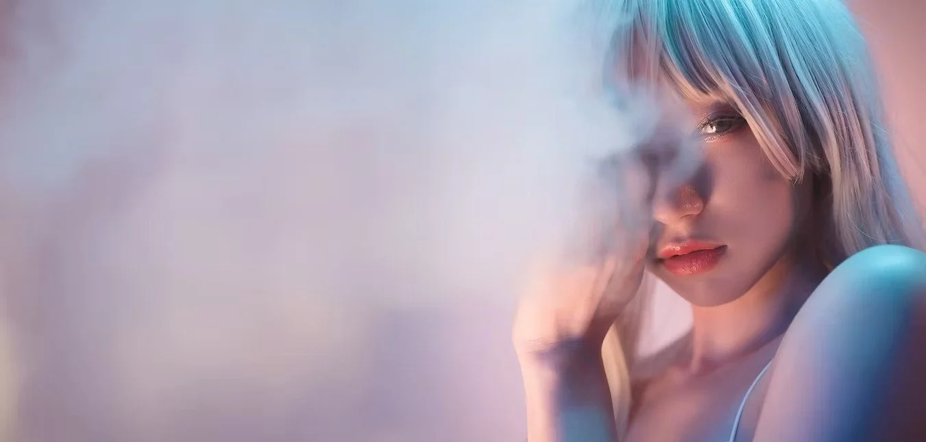 A woman with light-colored hair and makeup, looking at the camera through a cloud of smoke or mist with colorful lighting.