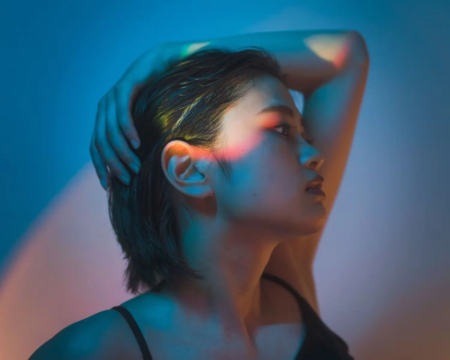 A woman with short hair poses with her hand behind her head against a colorful, gradient background.