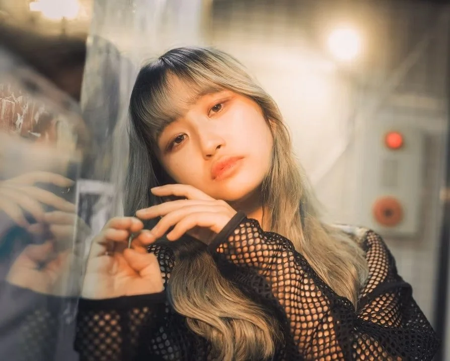 Young woman with long hair and bangs, wearing a black fishnet top, leaning against a reflective glass surface, with soft lighting and a slightly blurred background.