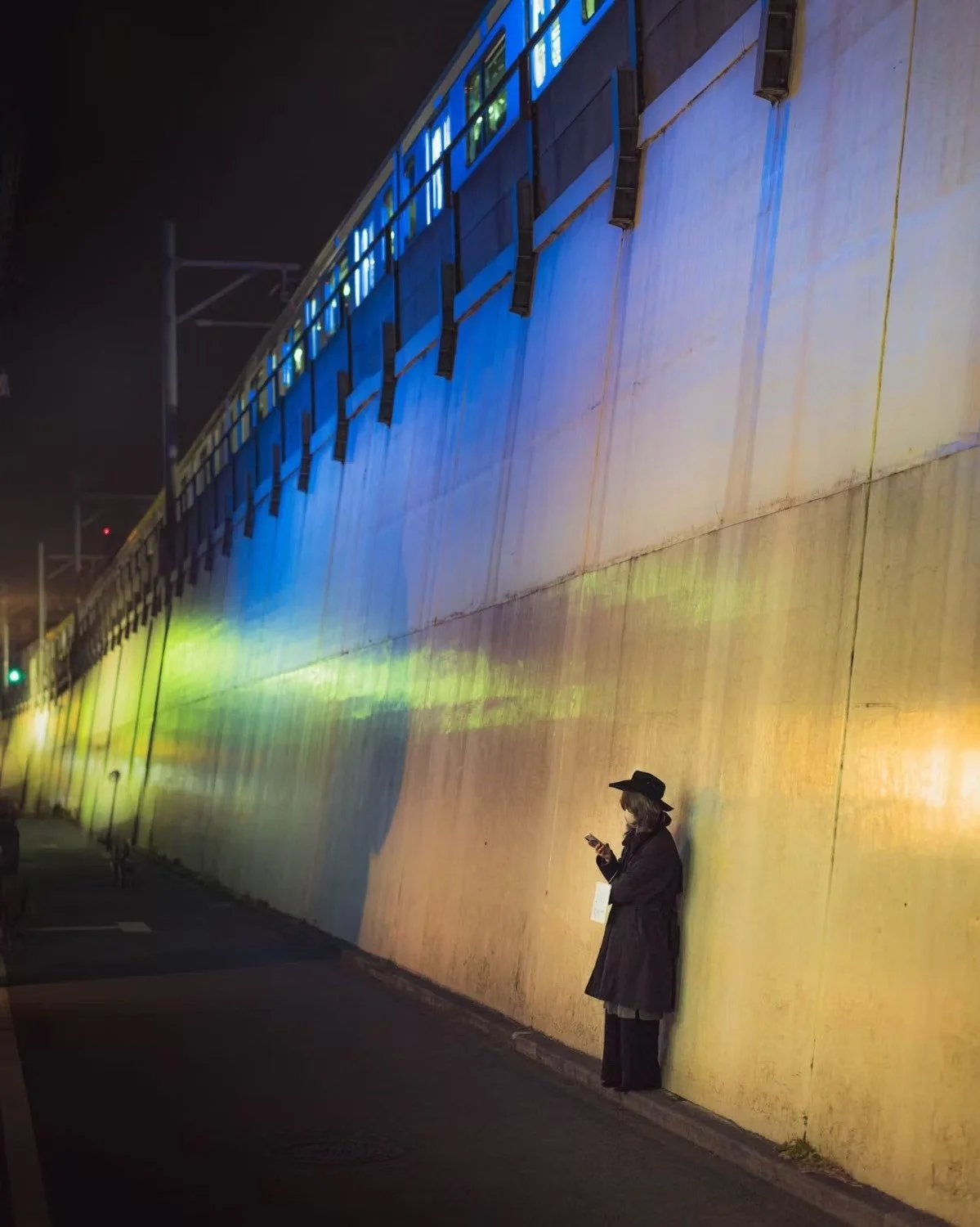 A woman standing against a brightly colored wall at night, looking at her phone. The wall is illuminated with blue, yellow, and green lights, creating a vibrant backdrop. She is wearing a long coat, a wide-brimmed hat, and a face mask.