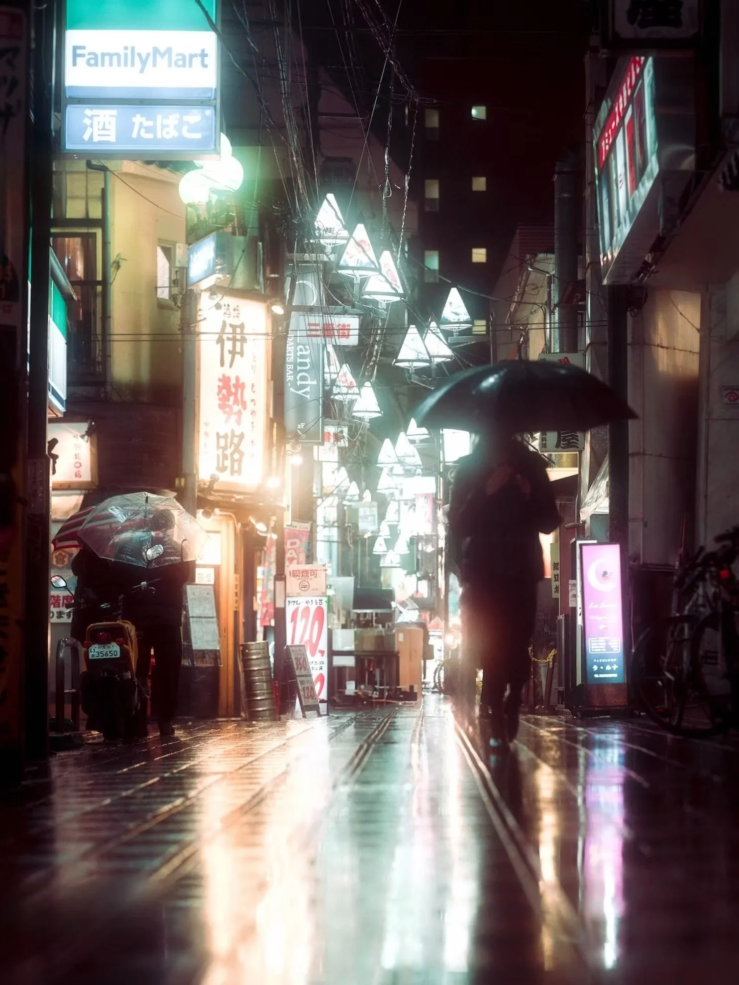 Nighttime street scene with illuminated signs in Japanese, umbrellas held by pedestrians, wet pavement reflecting lights, and overhead electrical cables.