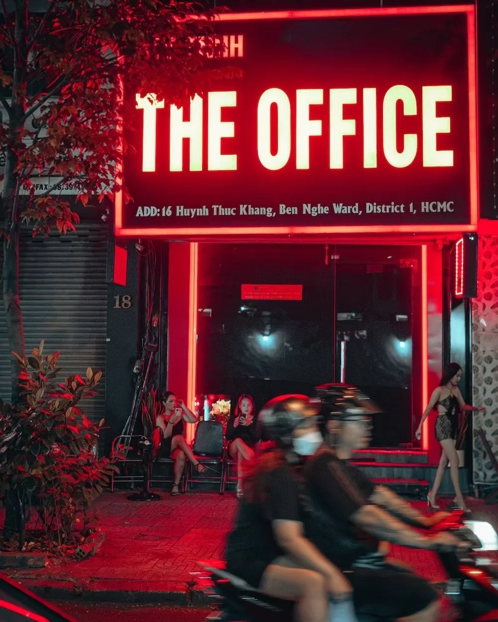 Nighttime city street scene with a bright red neon sign reading 'THE OFFICE' above the entrance. Three women sit on chairs outside, one wearing black, another in dress, and the third on her phone. Two women in black dresses stand nearby. Two motorbik