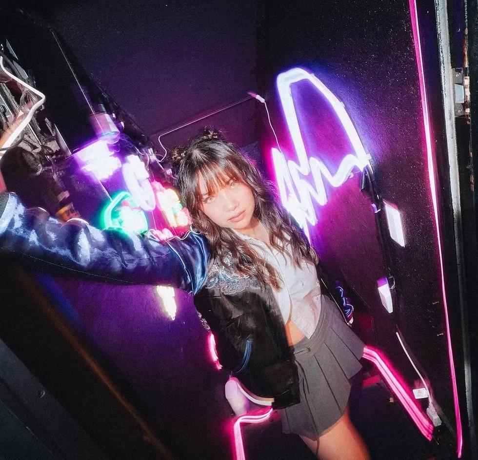 A young woman with wavy hair and bangs taking a selfie in front of a black wall with neon lights that spell out 'LOVE.' She is wearing a varsity jacket, a white top, and a pleated gray skirt, with neon lighting reflecting off her clothes and the wall.