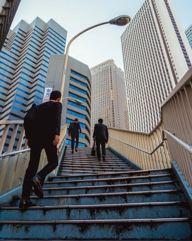 Three salarymen in Tokyo Shinjuku in suits walking up outdoor stairs in a city with tall skyscrapers and a streetlight overhead shot by photographer Deniz Demir.