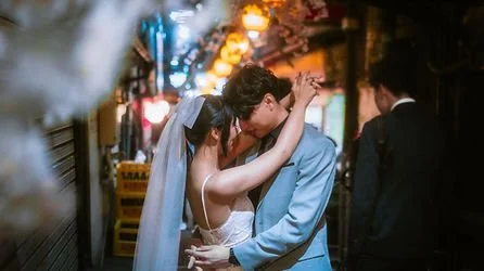 A couple in wedding attire embracing and about to kiss on a city street at night, with blurred background lights and people.