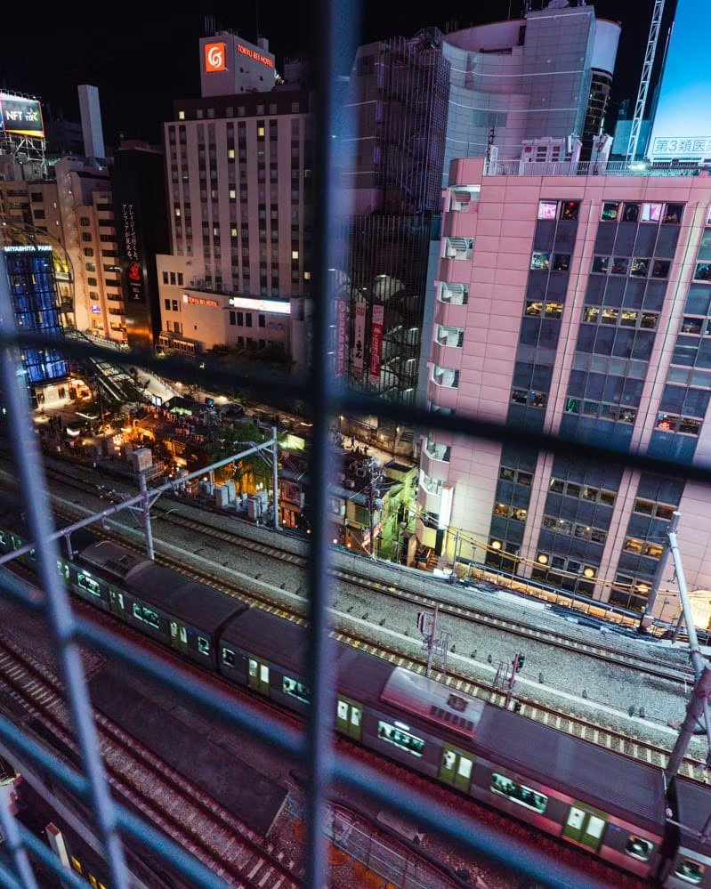 Cityscape at night seen through a grid barrier showing tall buildings, a train on elevated tracks, and various illuminated signs in a metropolitan area.