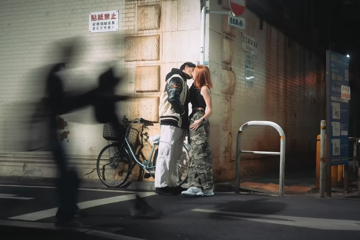 A couple shares a kiss in a dimly lit Tokyo alley with a bicycle in the background. The scene is illuminated by streetlights, and a blurred figure passes by in the foreground.