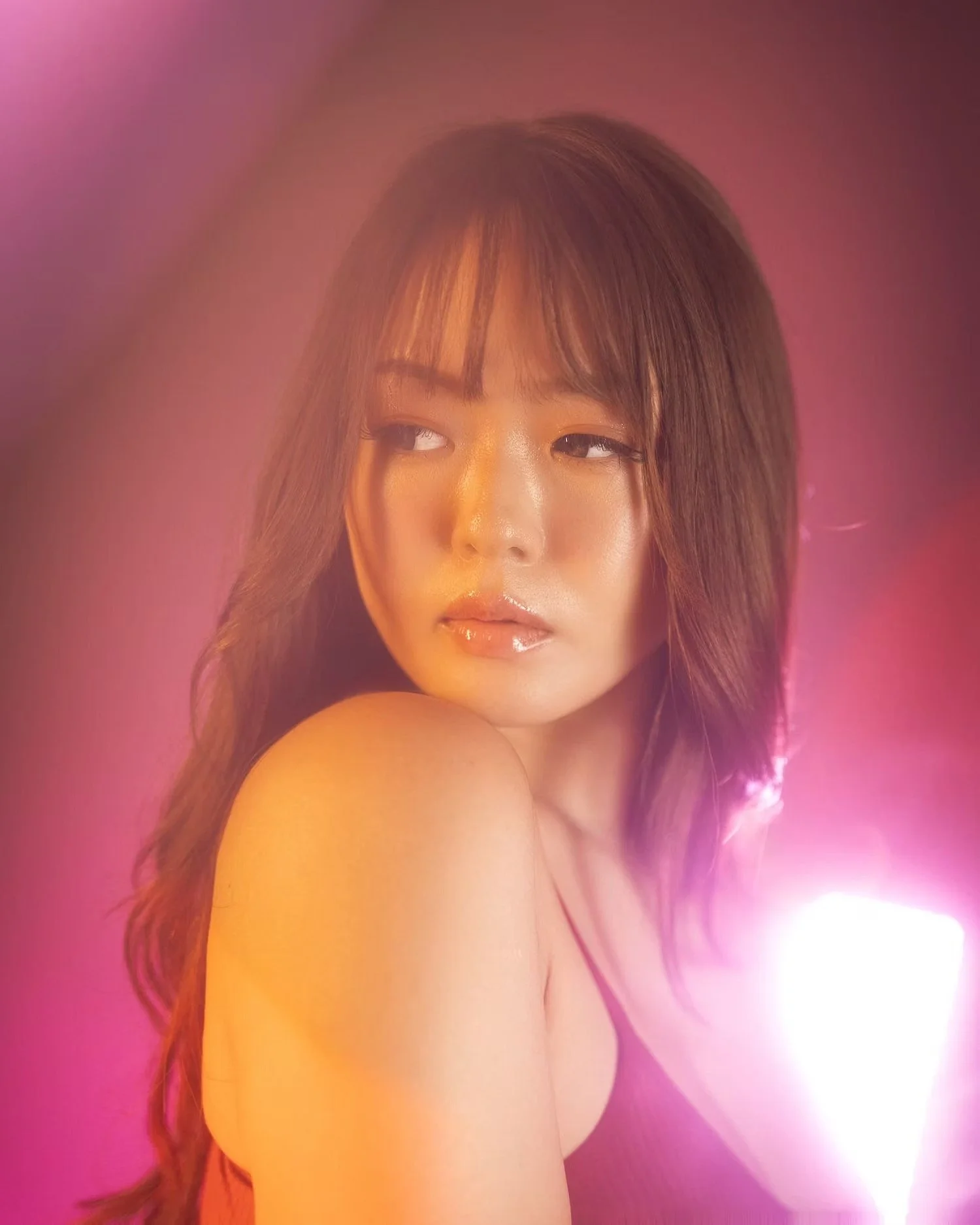 A woman with long brown hair and makeup posed against a pink background with light effects.