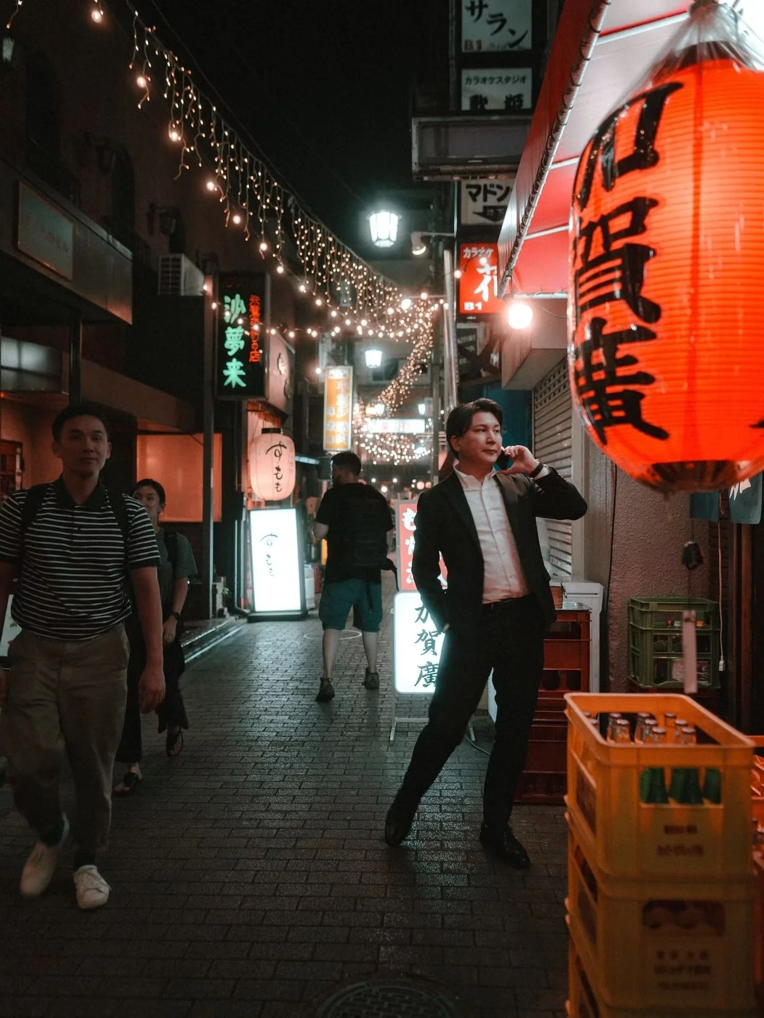 Night scene of a busy street in Japan with hanging lanterns and string lights, several people walking, illuminated signs in Japanese, and a large red lantern on the right side.