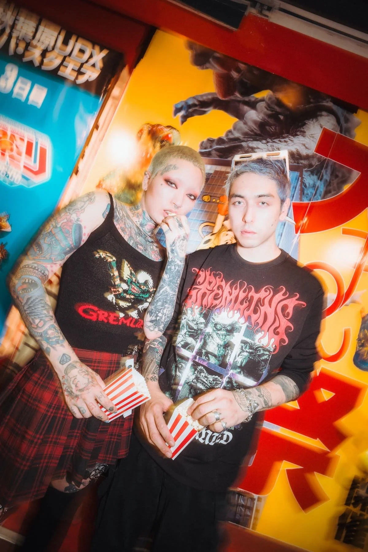 A woman with tattoos and green hair and a man with dyed gray hair are standing together holding popcorn, inside an arcade or entertainment venue with colorful artwork and a blue and red wall background.