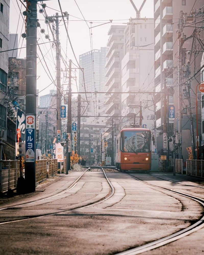 A street scene with a tram approaching on tracks, surrounded by tall buildings and utility poles with wires, in a city setting.