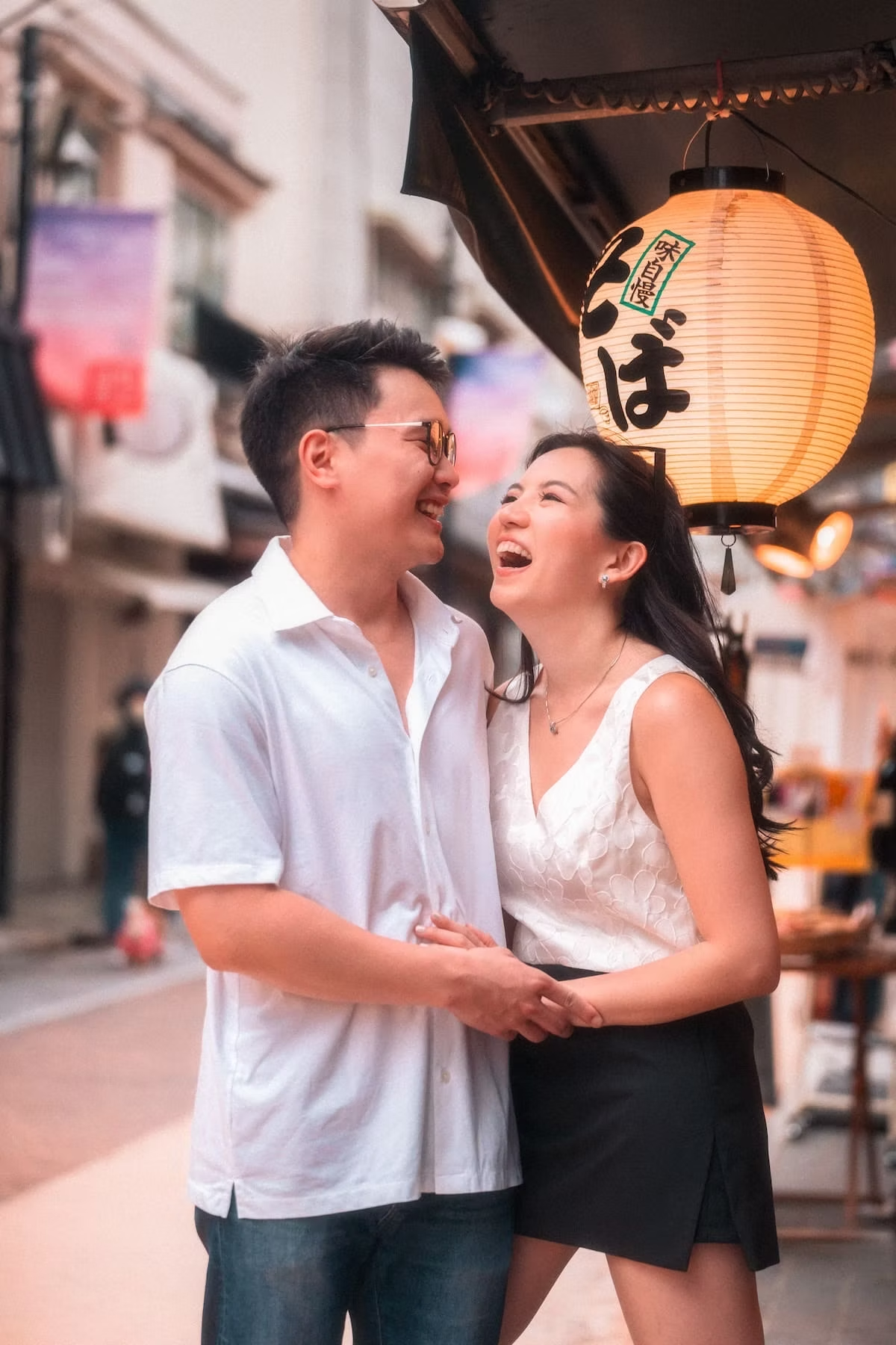 A couple stands laughing and holding hands in a Tokyo alley lit by traditional lanterns, with the soft glow of the lanterns enhancing the romantic atmosphere.