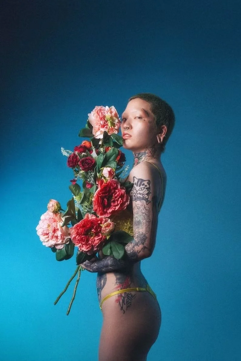 A woman stands with her back slightly turned, holding a large bouquet of vibrant flowers. The rich lighting and her confident stance convey strength and beauty, with the flowers adding a touch of femininity and elegance.