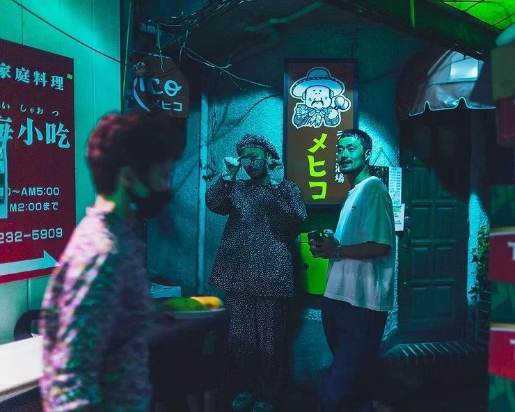 Two men standing outside a restaurant in Kabukicho, one wearing a polka-dot outfit and gloves, captured with colorful neon lights around them.