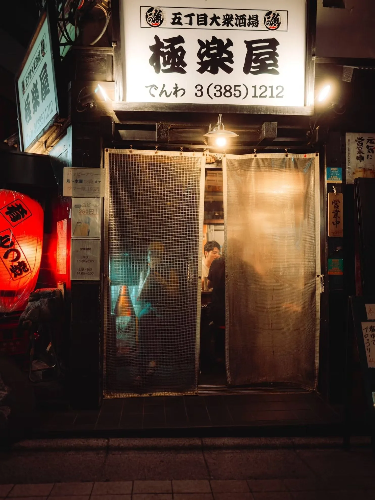 A Japanese restaurant or bar entrance with illuminated signs, traditional red paper lantern, and beige fabric curtains, revealing a man inside.