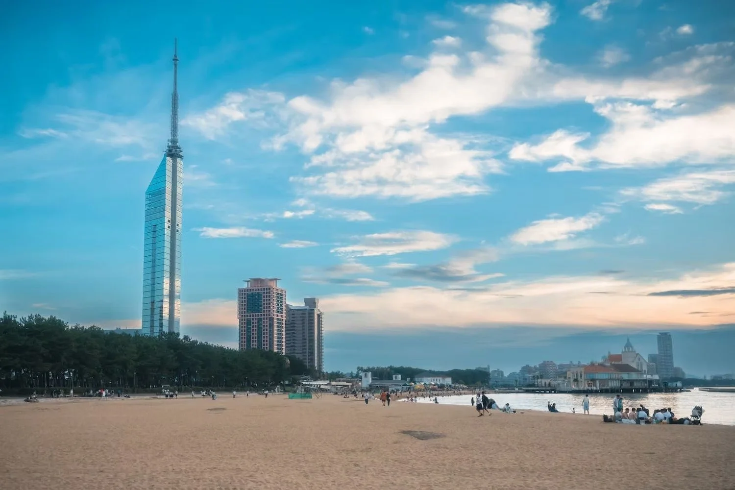 A beach in Japan with people sitting and walking, high-rise buildings including a tall skyscraper, greenery along the shoreline, and a partly cloudy sky at sunset.