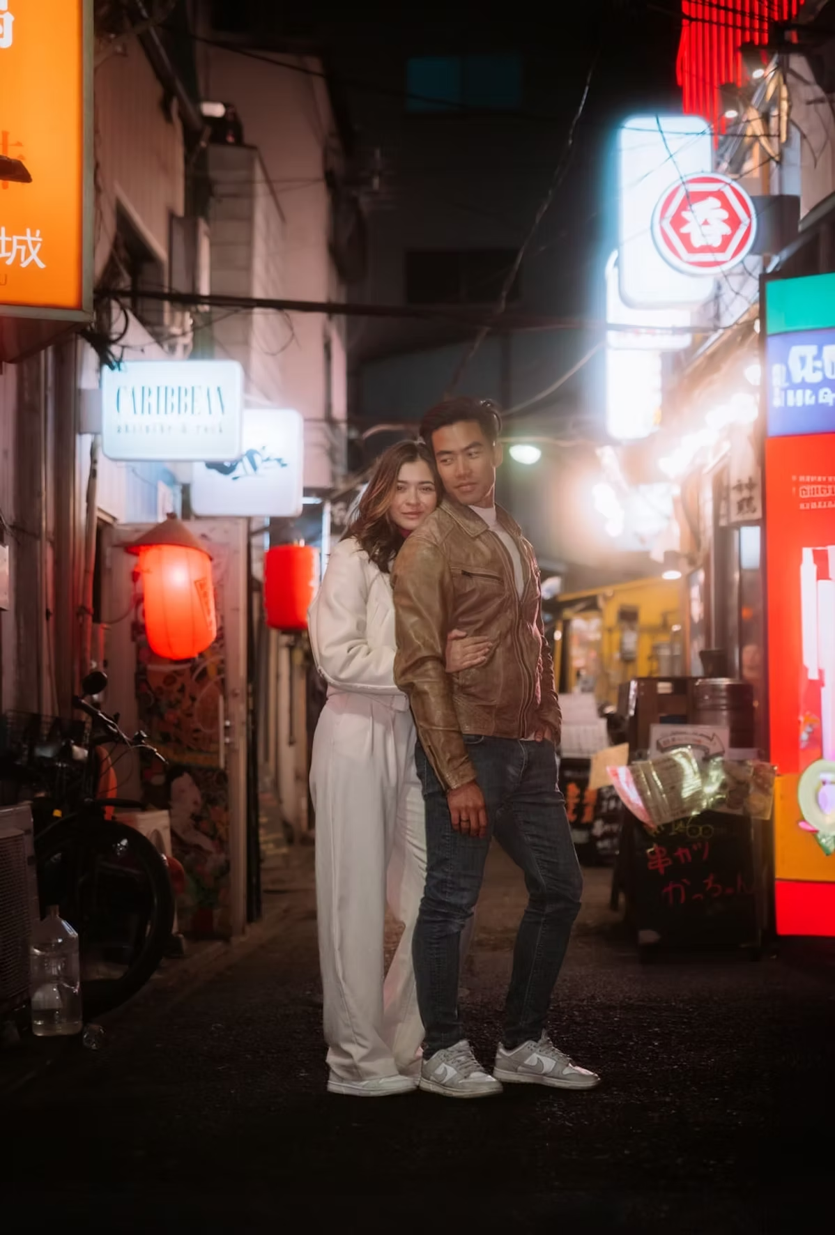 A couple stands together in a quiet Tokyo alley, posing for a photo. The woman wears a white sweater and the man is dressed casually. The alley is lit by soft streetlights, with vibrant neon signs visible in the background.