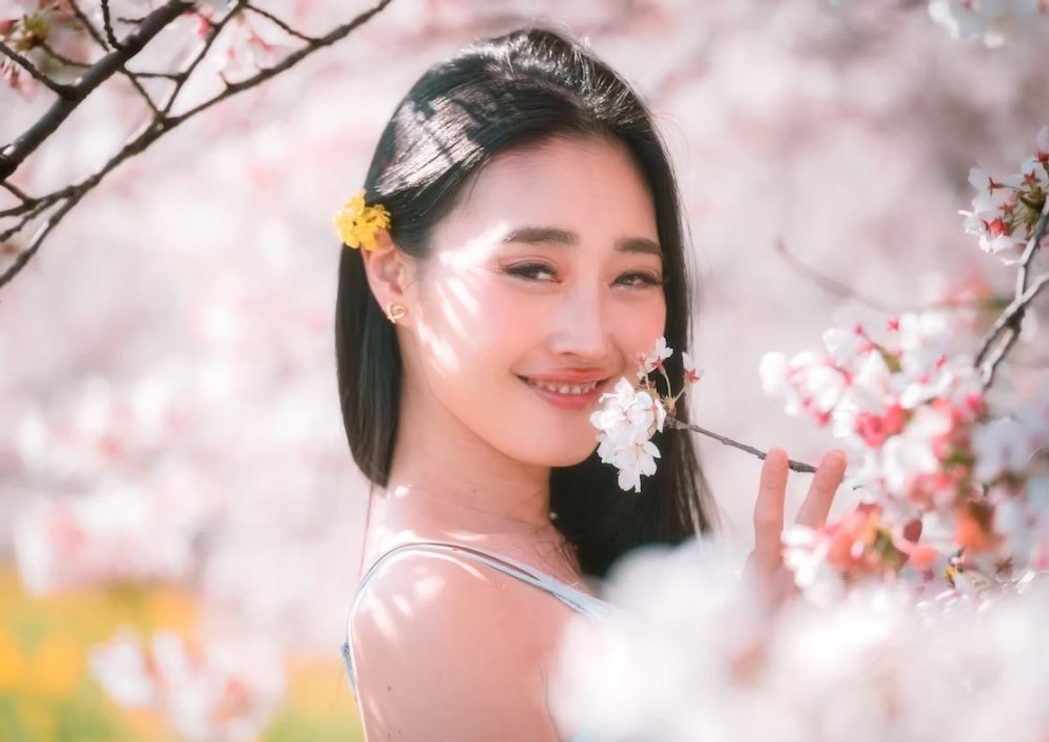 A woman smiles at the camera while holding a cherry blossom branch. A small yellow flower is tucked behind her ear, and she stands surrounded by blooming cherry blossoms, creating a peaceful, springtime vibe.