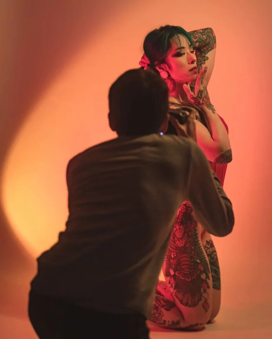 A photographer captures the moment as a model in a colorful outfit poses under warm lighting. The image shows the behind-the-scenes process of creating a dramatic photo, with the photographer’s figure subtly visible in the shot.