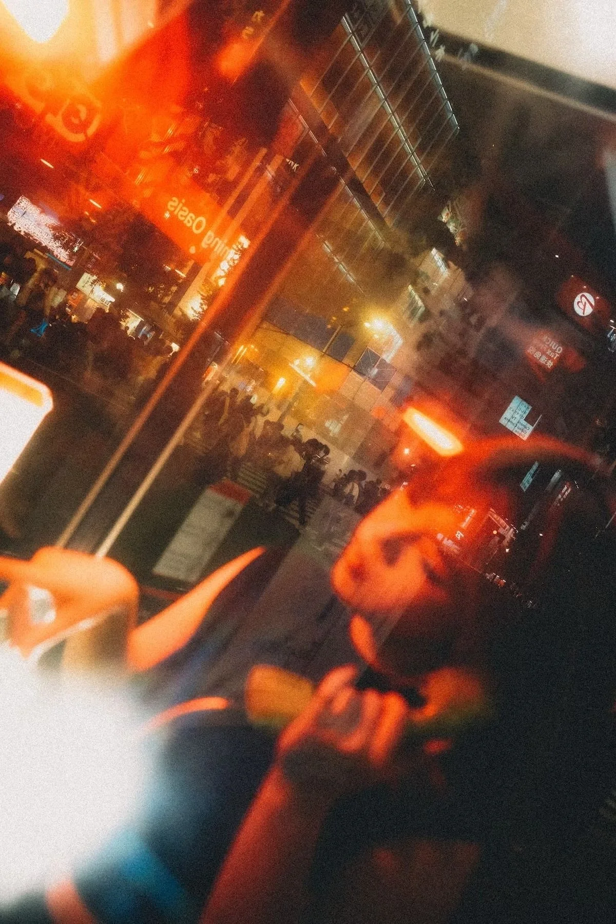 Night scene viewed through reflective glass showing blurry lights, a crowd of people, and illuminated signs in an urban area. Shot by Tokyo portrait photographer Deniz Demir.