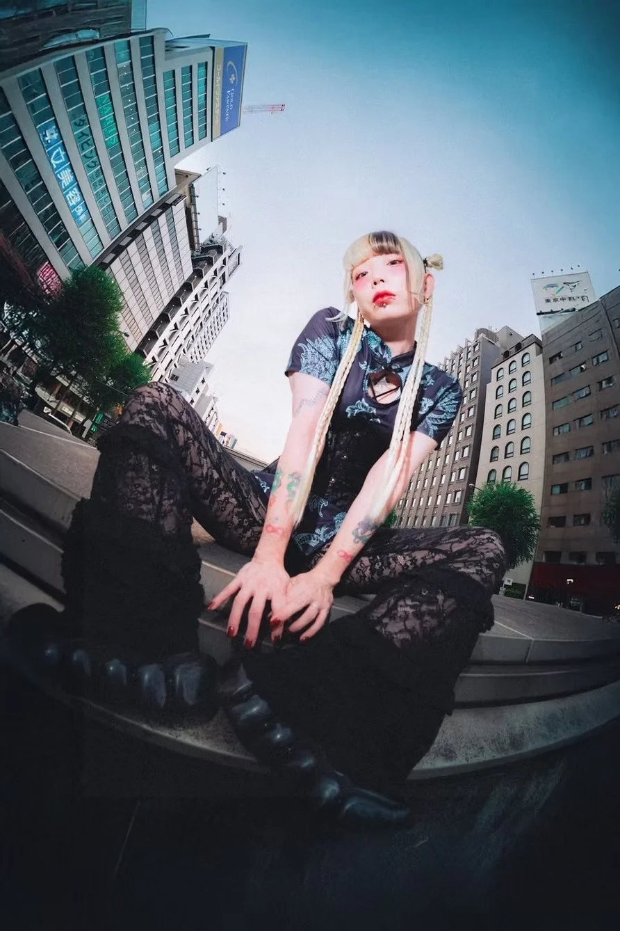 A young woman with long blonde hair, tattoos, and piercings, sitting on a city sidewalk with tall buildings in the background. She wears a dark floral dress, lace tights, and black boots, posing with her legs spread apart and looking at the camera. W