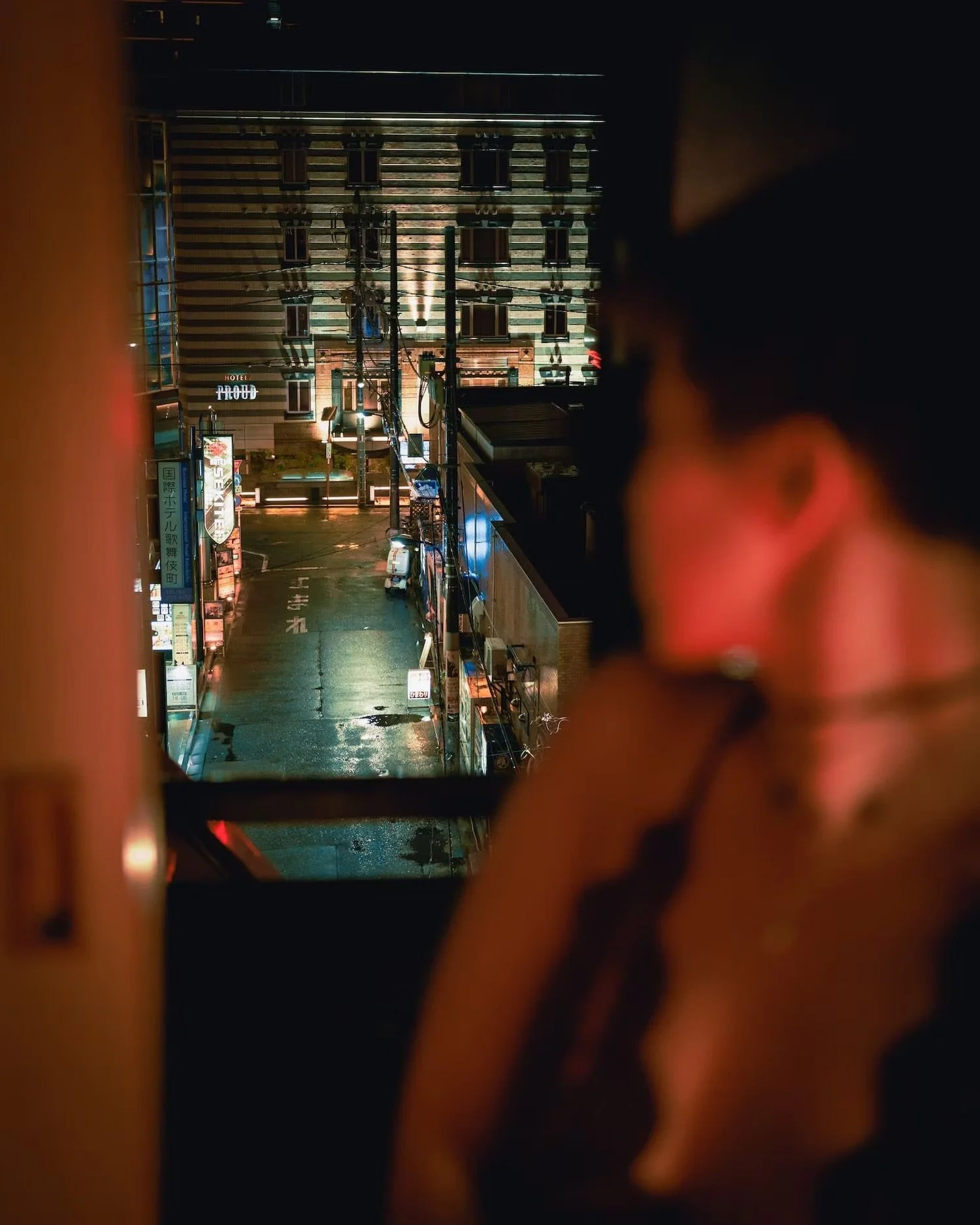 Nighttime view of a street with a wet road, illuminated signs in Japanese, and a building in the background with a striped pattern facade. The photo is taken through an opening or window with dark and blurry edges.