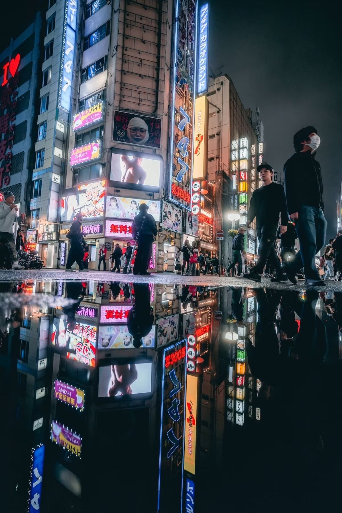 Night scene of a busy city street with neon signs and advertisements in Japanese, reflecting on a wet pavement, with pedestrians wearing masks walking among illuminated buildings. Kabukicho in front of Godzilla shot by Tokyo street photographer Deniz