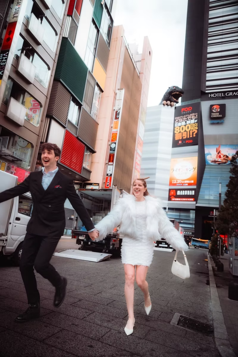 A couple in casual winter clothing walks hand in hand along a busy street in Tokyo, with colorful buildings and advertisements surrounding them.