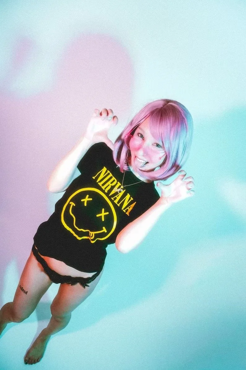 A woman with short pink hair wearing a Nirvana t-shirt stands against a blue background. She strikes a playful pose, with one hand on her hip and a relaxed expression on her face, adding to the carefree and youthful vibe of the image.