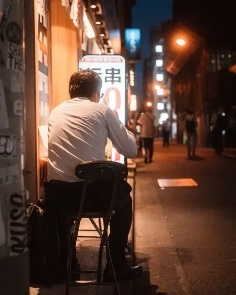 A person seated at a street-side table in Tokyo, illuminated by neon lights. Captured during the Tekumi Photowalks street photography workshop.