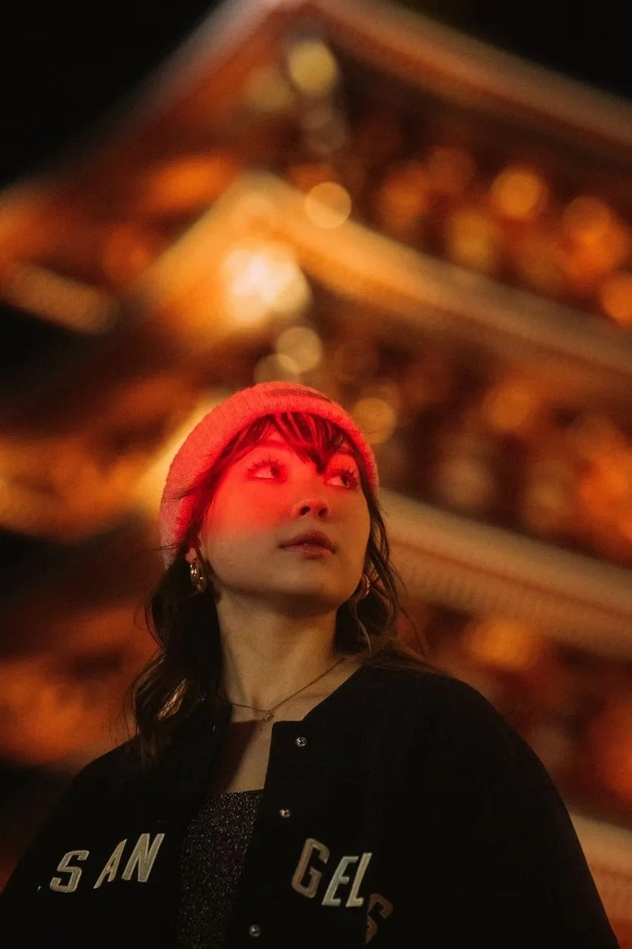 A young woman with dark hair, wearing a red beanie, stands in front of a brightly lit, blurred building at night, illuminated by red light in Asakusa, Tokyo.