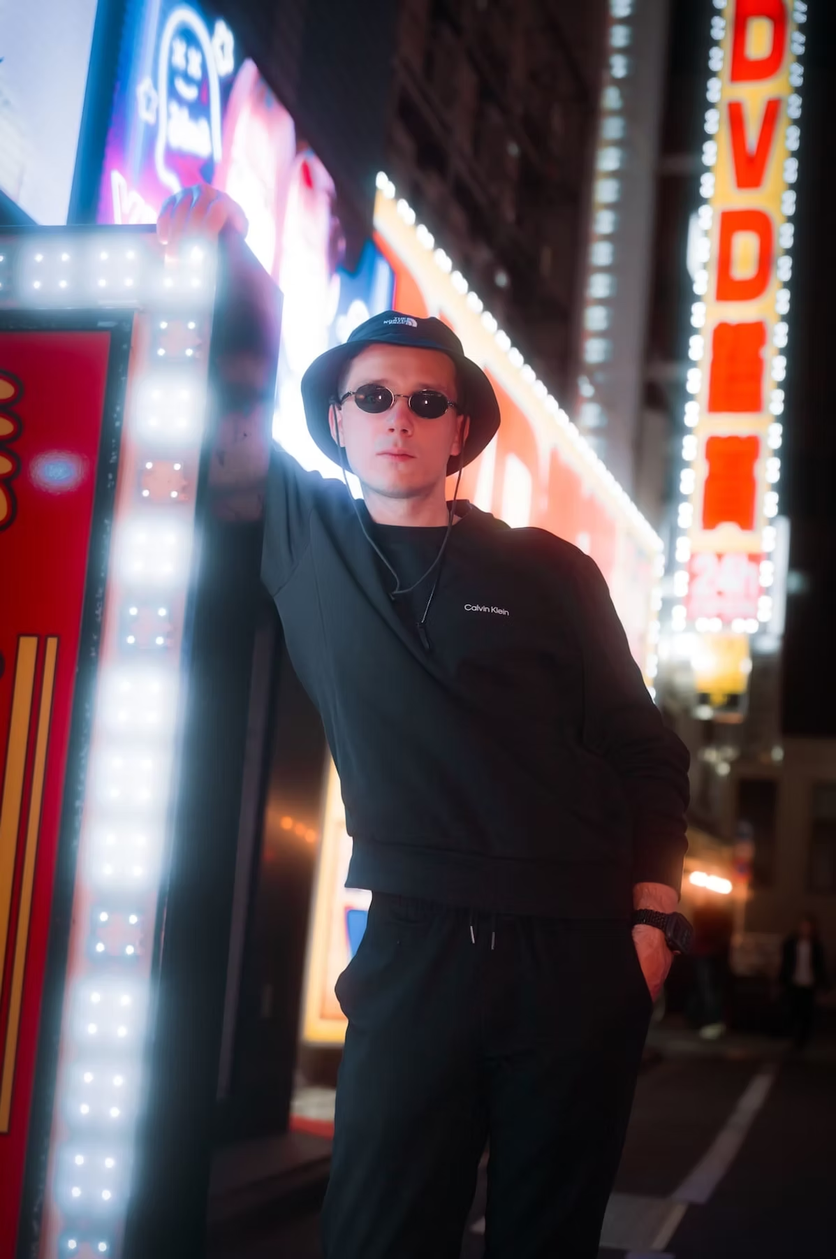 A man in a black hoodie, pants, and a hat stands casually with one hand on a glowing signpost in the vibrant streets of Tokyo at night.