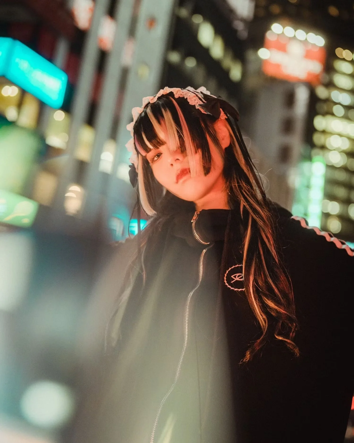 A woman with black and blonde hair, wearing a black hoodie with a white logo, standing in an urban, illuminated cityscape at night with blurred colorful lights in the background.