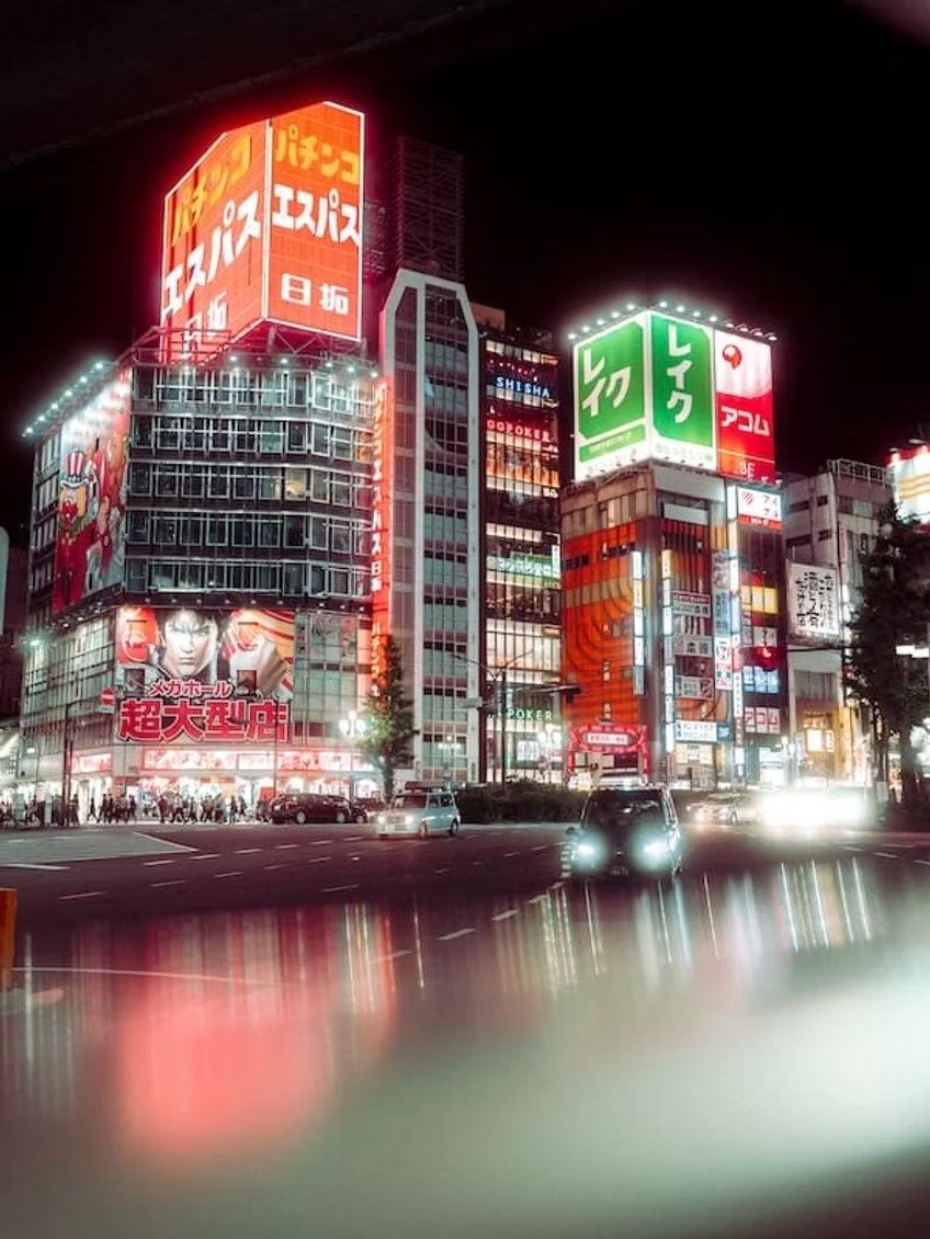 Nighttime city street in Japan with illuminated billboards and signs in Japanese, cars on the road, and a building with red and green advertisements.