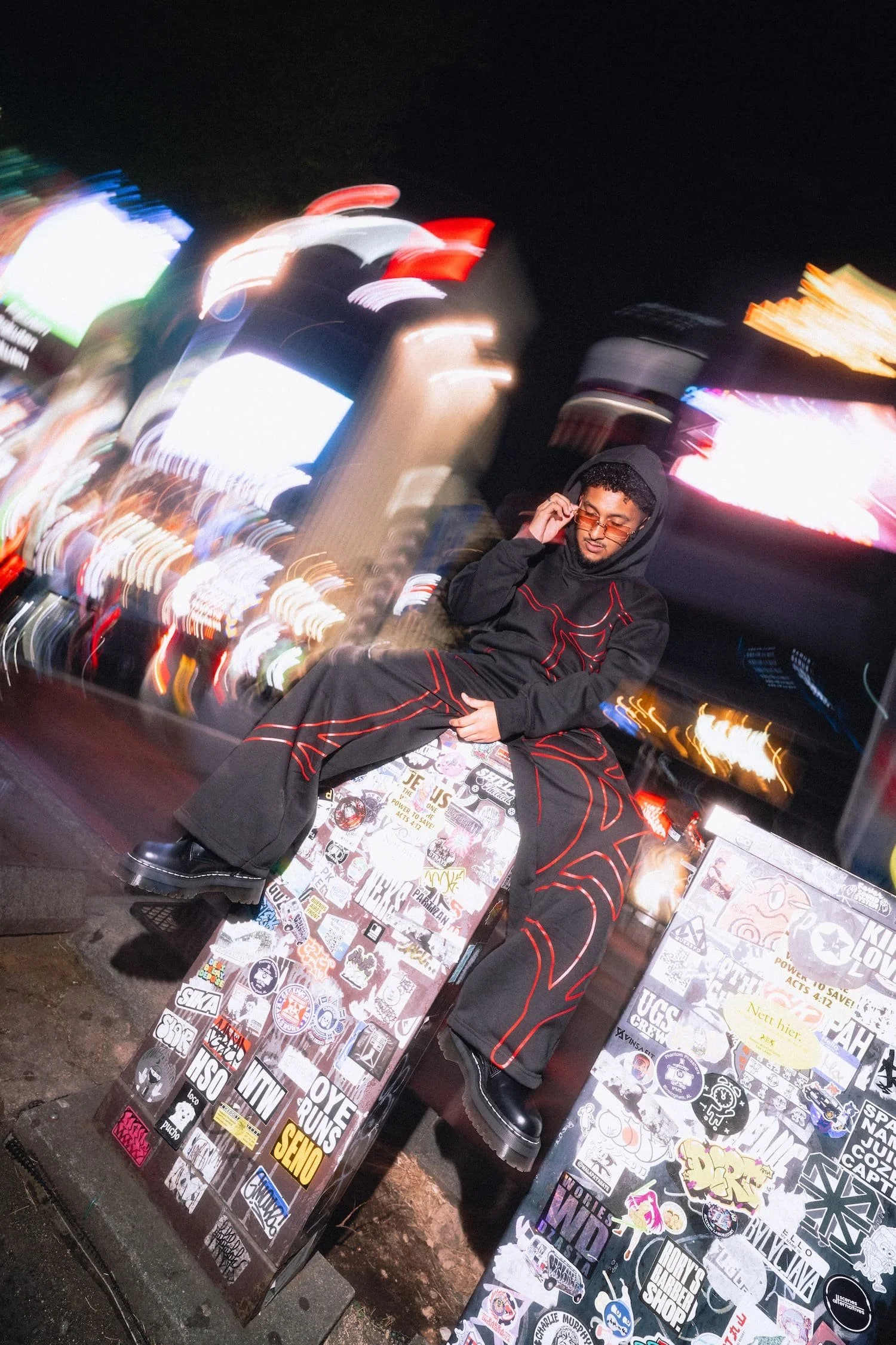 Man sitting at a city street corner at night, talking on a cell phone, with various street signs and neon lights blurred in the background, and stickers on the tables in front of him with Shibuya Crossing in the background. Shot on wide angle flash.