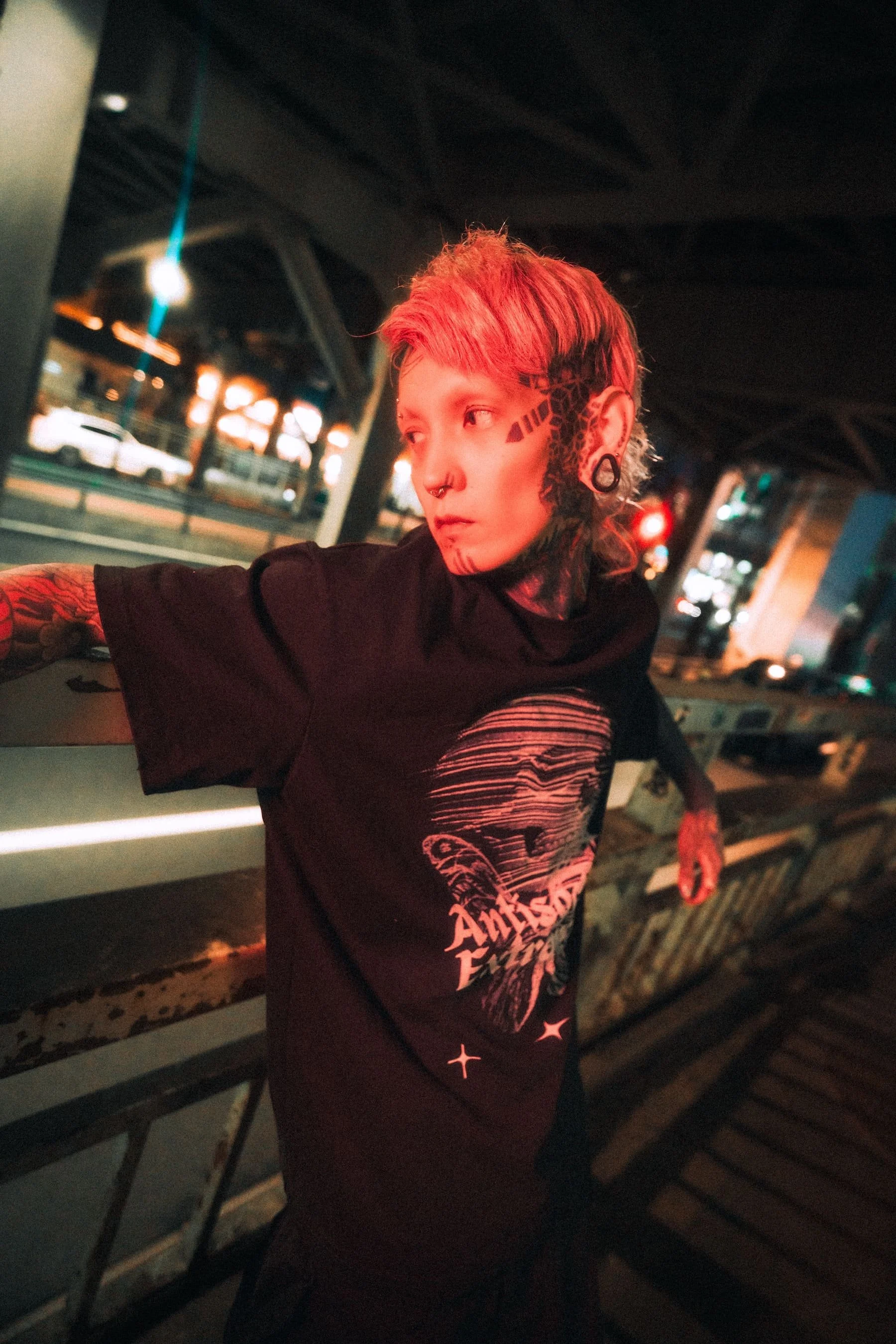 A person with short, light-colored hair and tattoos on their face and arm, wearing a black T-shirt with white text, leaning against a metal railing at night with city lights in the background.