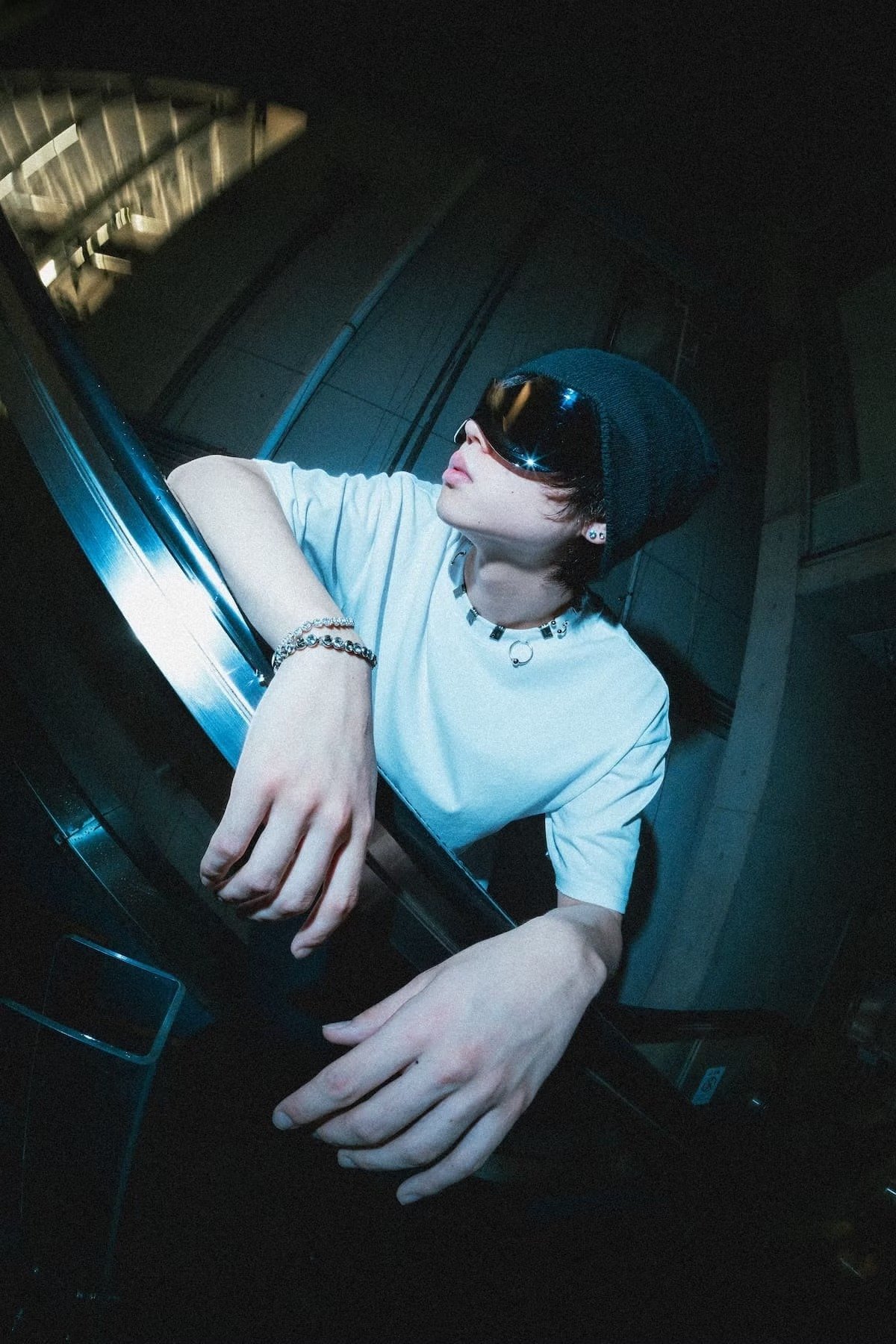 A model wearing a white Fragile Base t-shirt, beanie, silver jewelry, and futuristic sunglasses leans over a reflective metal railing at night. Harsh flash lighting and deep shadows emphasize Japanese street fashion and cyber-influenced styling. Shot