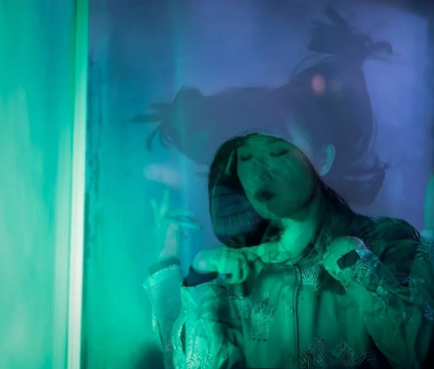 A woman with dark hair and makeup, reflected in a colorful, abstract blue-green surface, with a contemplative expression and closed eyes, creating a layered, artistic effect. Shot by Tokyo portrait photographer Deniz Demir.