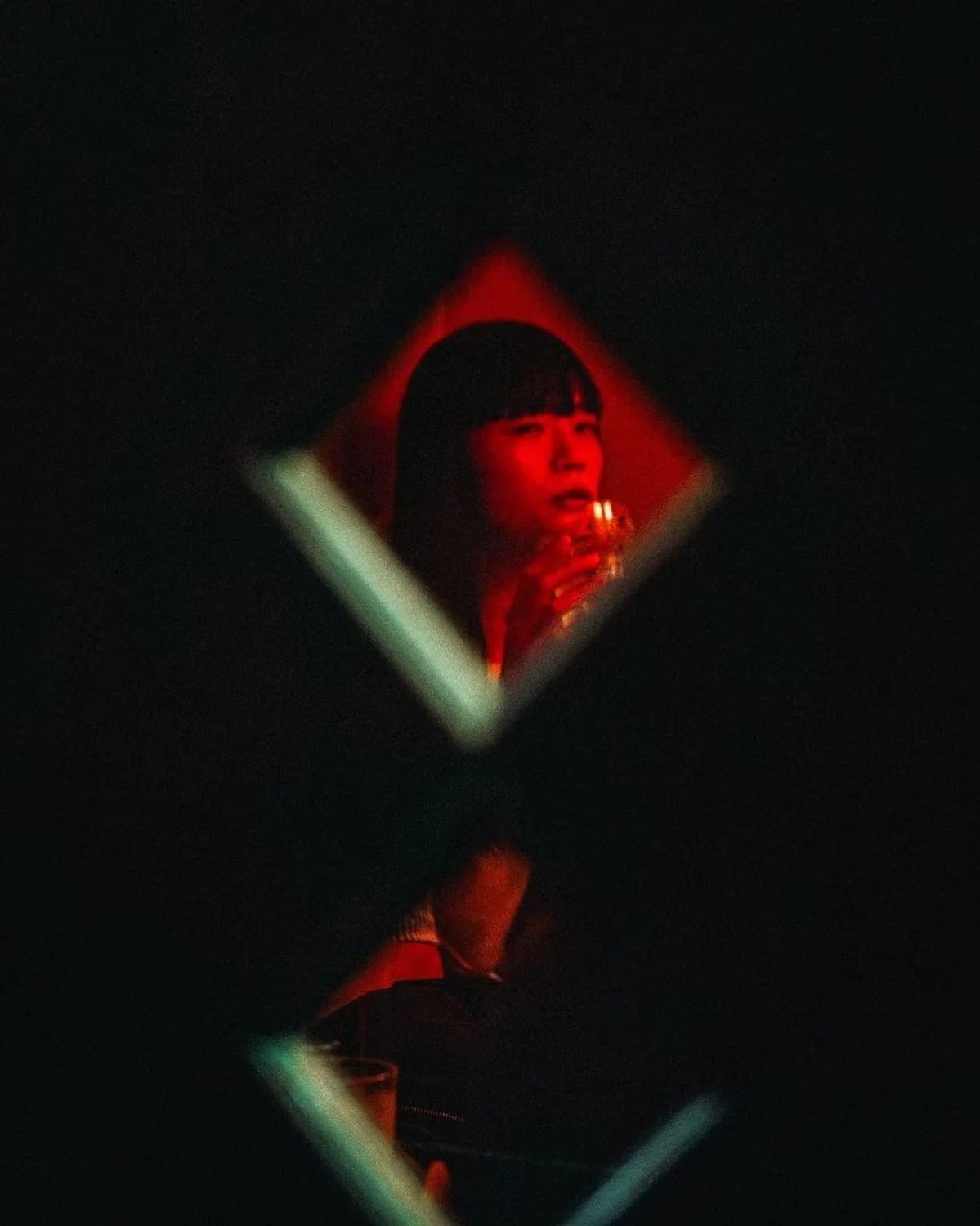 A Japanese woman with short hair and bangs holds a drink, illuminated by red light, reflected in a small square mirror with a dark background.