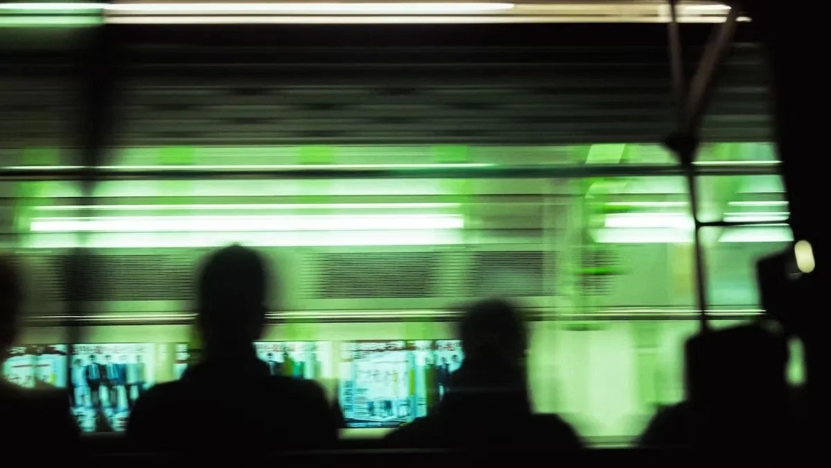 Street photography of people sitting on a Tokyo train with a green storefront visible outside the window.