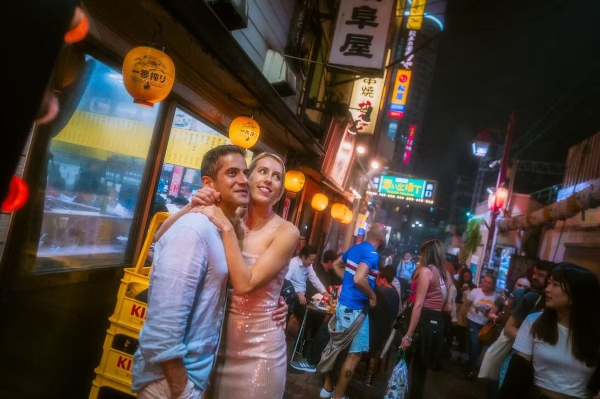 A couple poses together in a narrow Tokyo street lit by yellow lanterns. The vibrant, bustling street is filled with people enjoying the nightlife, while the couple smiles at each other, creating a romantic atmosphere.