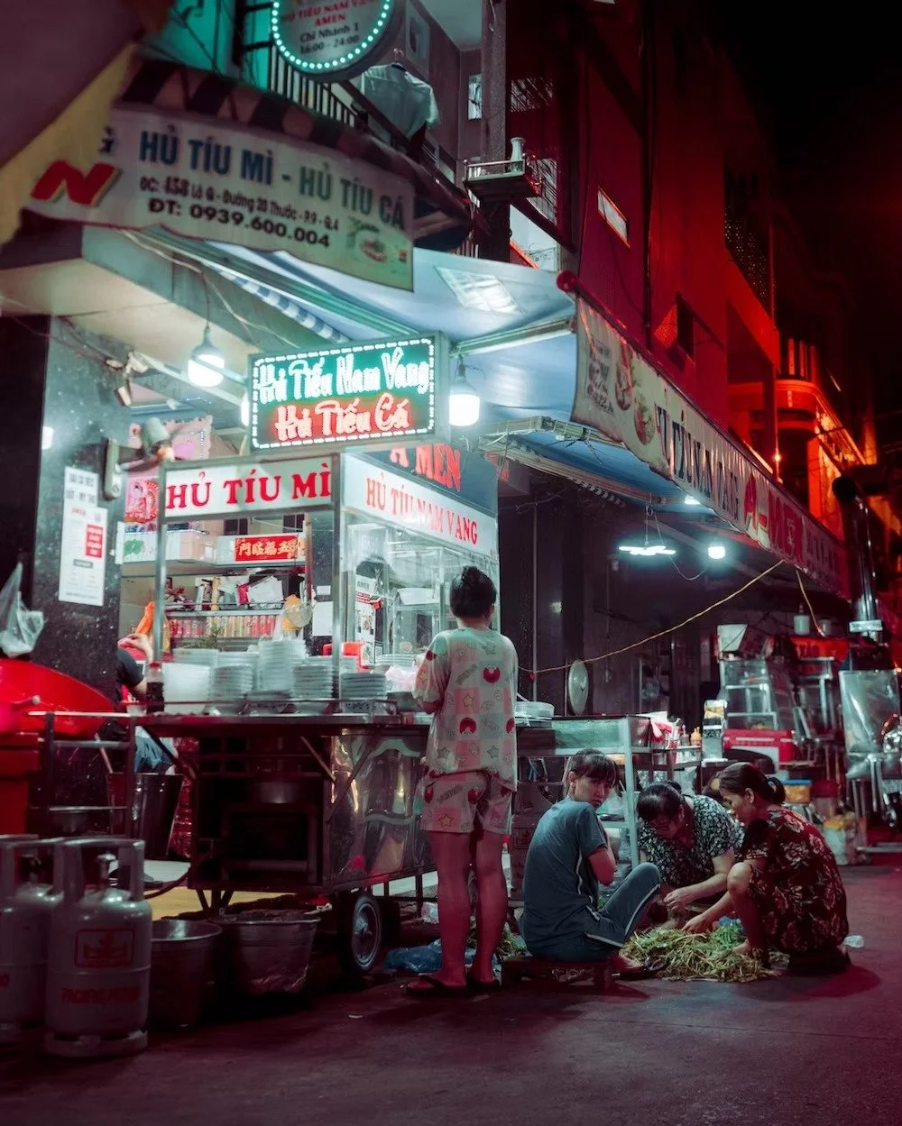A street food vendor stall at night selling Vietnamese dishes, with three women seated on the ground preparing vegetables, and a woman in pajamas standing nearby. Neon signs and street lights illuminate the scene. 