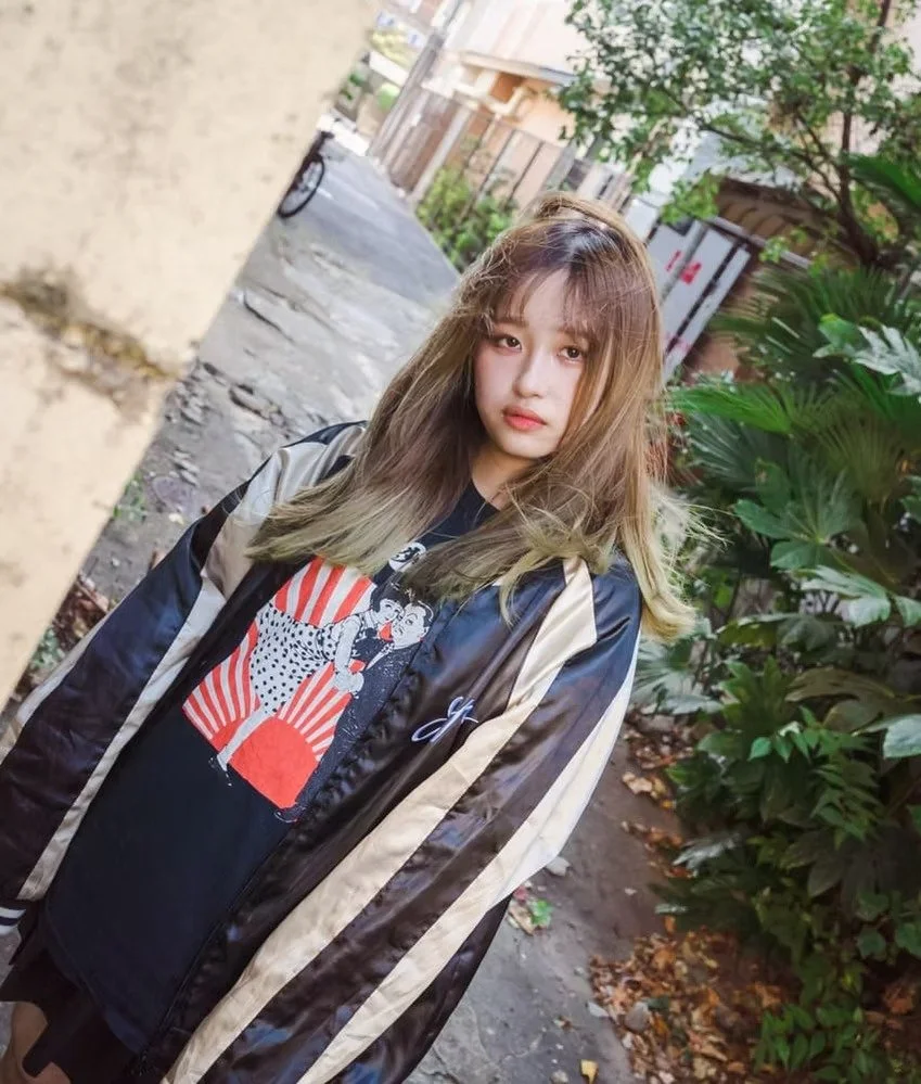 A young woman with long, light brown hair standing outdoors on a sidewalk with green plants nearby. She is wearing a black t-shirt with a colorful graphic and a shiny, striped jacket. The background shows a building and a tree.