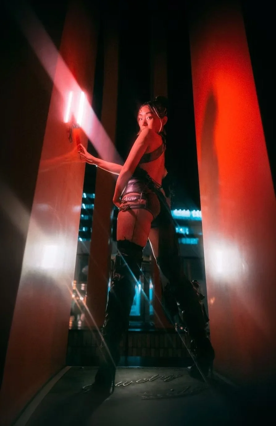 A woman in black leather clothing posing in a narrow hallway with red neon lighting.